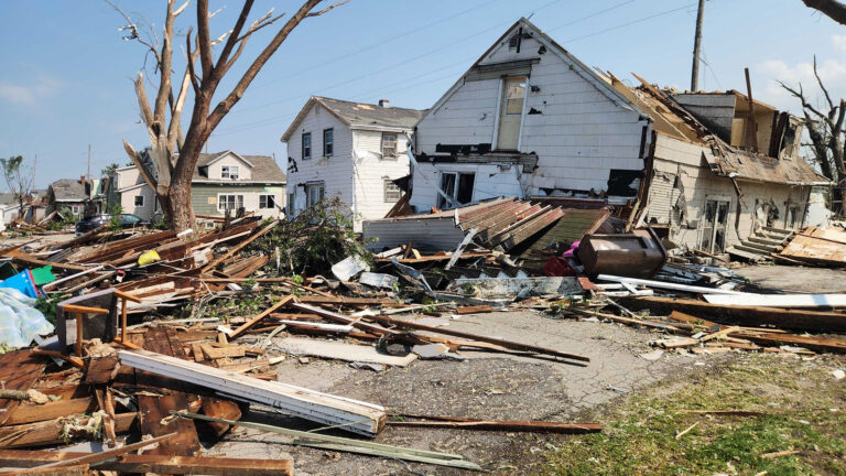 Siding, wall studs and other types of wood debris sits in piles on the ground among trees stripped of leaves and small branches and in front of a damaged two-story house with no roof, with other damaged houses in the background.