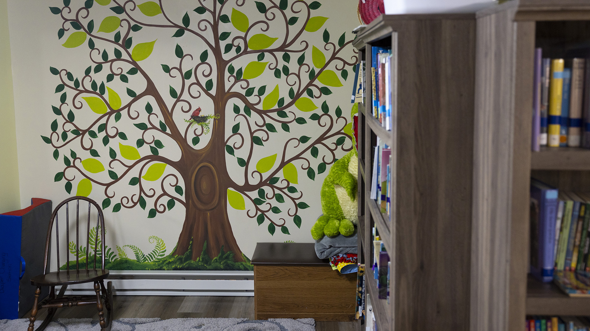 A small rocking chair sits in front of a wall painting of a stylized tree with a cardinal and its nest at its center, with a small wood box on the floor next to multiple bookshelves filled with books extending into the foreground.