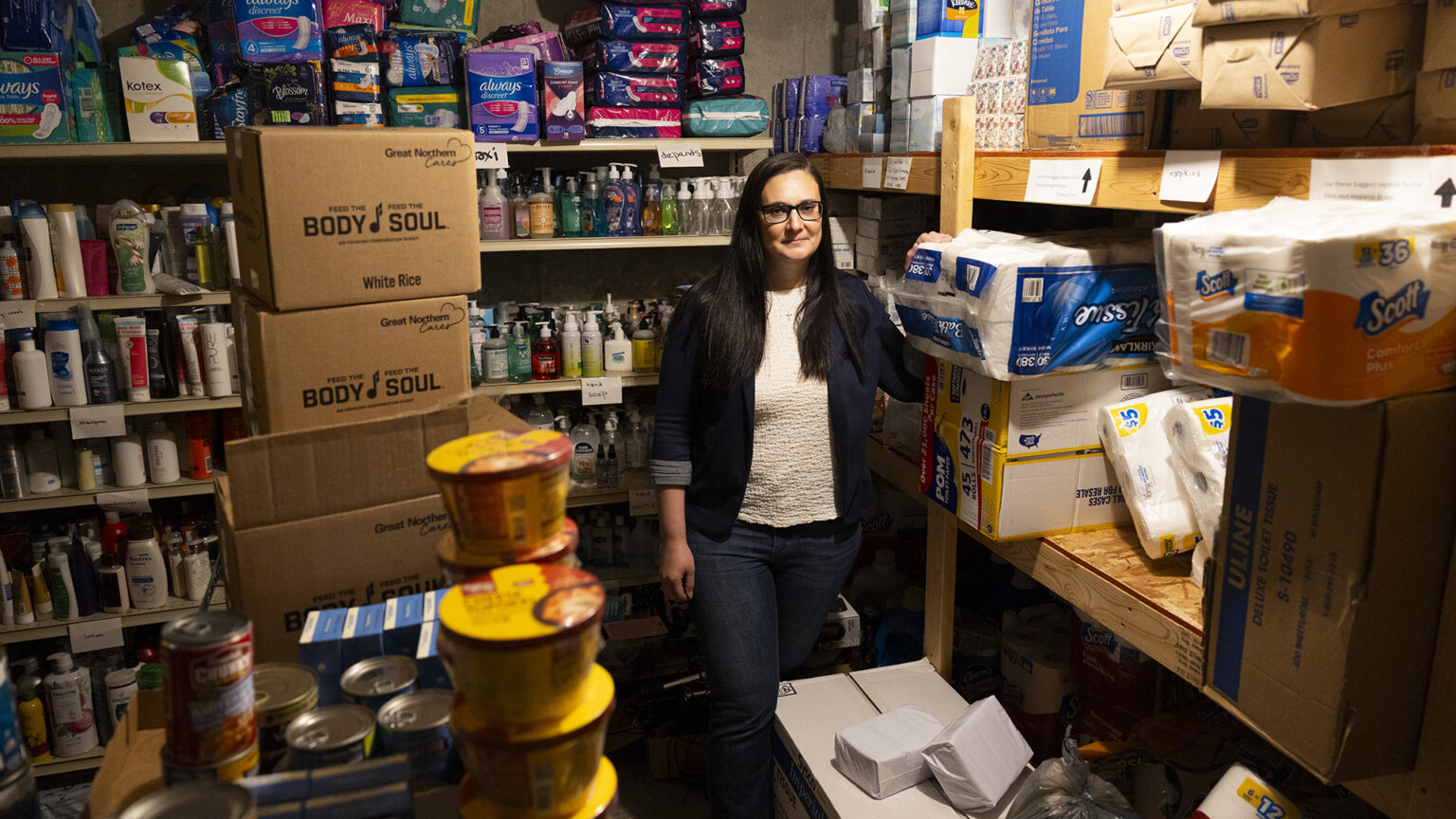 Mindy Lohff poses for a portrait while standing in a storage room with wood shelves filled with cases of toilet paper, facial tissue, sanitary napkins, soaps, shampoos and other personal toiletry items, with stacks of canned and dry food items in the foreground.