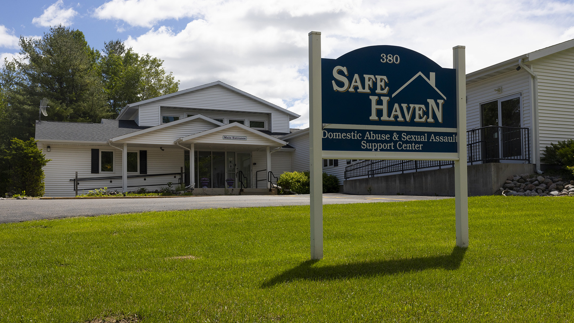 A wood sign with the address number 380, the words "Safe Haven" integrated into the logo of a house with a chimney, and the words "Domestic Abuse & Sexual Assault Support Center" is mounted between two wood poles in a lawn in front of a building with multiple wings, with two pediments framing one entrance and a concrete ramp with a metal railing leading to an accessible entrance, with trees in the background.