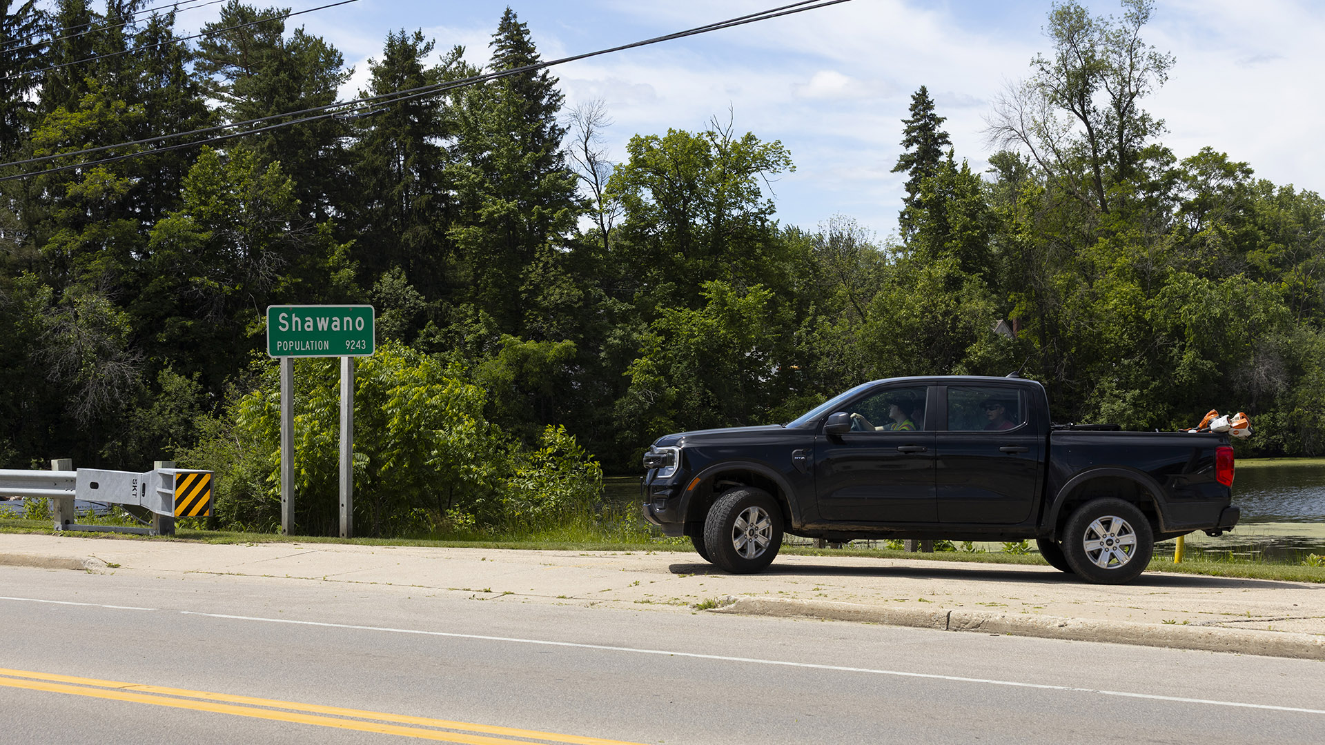 A crew-cab pickup truck drives along a driveway toward a road, with a municipal sign reading "Shawano" and "Population 9243" next to the end of a guardrail, with a river and trees in the background.