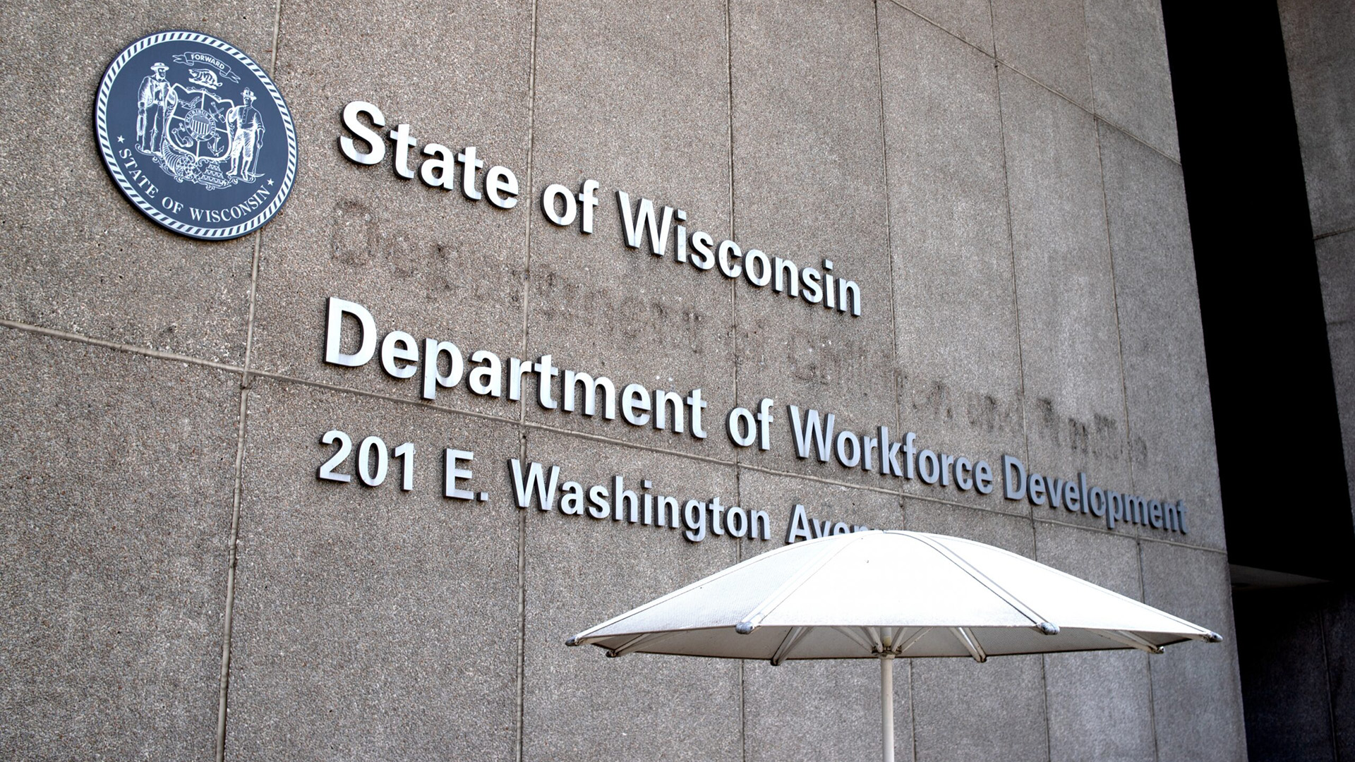 An outdoor patio umbrella stands in front of a building with poured concrete masonry, with the Wisconsin state seal affixed to the wall next to a letter sign reading "State of Wisconsin," "Department of Workforce Development," and an address, with weathering visible where a different letter sign was previously affixed.