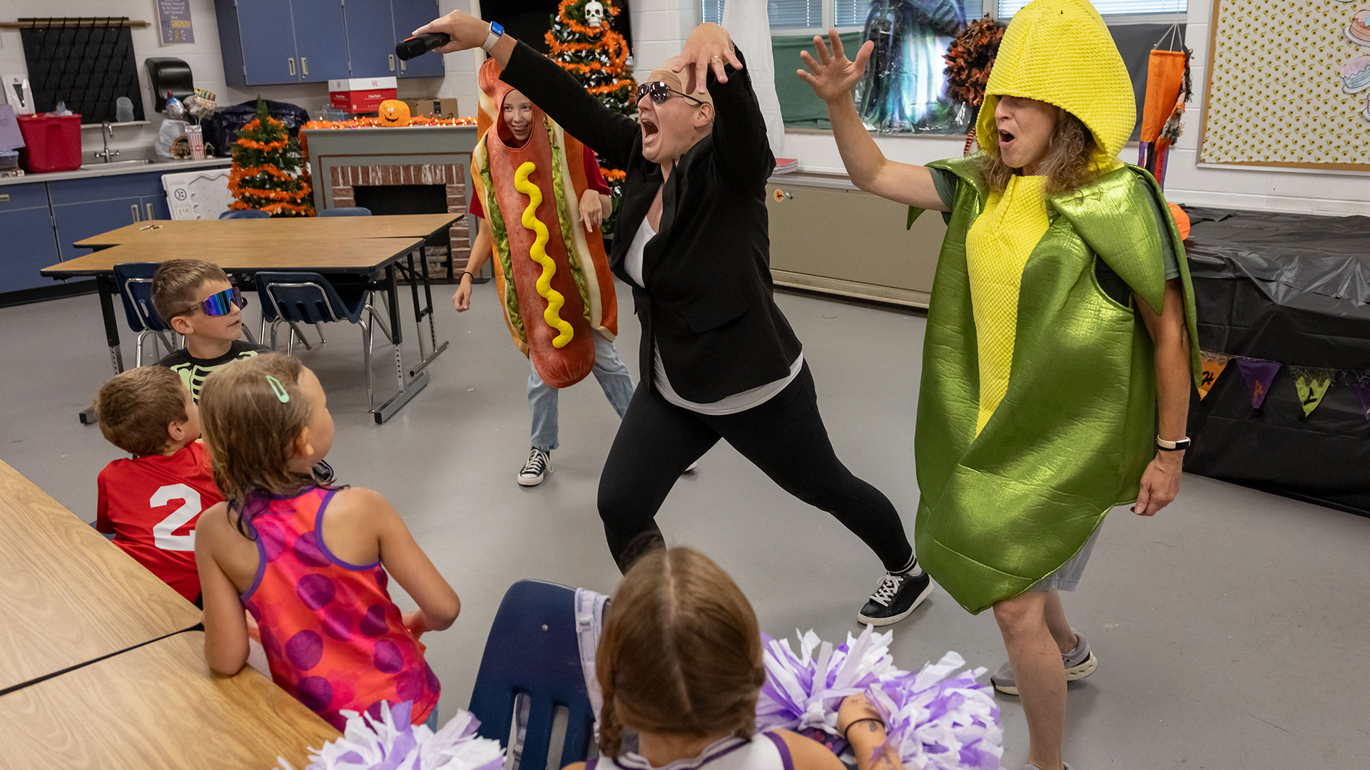 Stephanie Breitenbach holds both arms in the air in a dance pose with two other people, one in a hot dog costume and another in an ear of corn costume, in motion on either side of her, facing children seated in small stackable chairs next to low tables, in a room with a vinyl floor, additional low tables, wall cabinets and a sink, and a window.