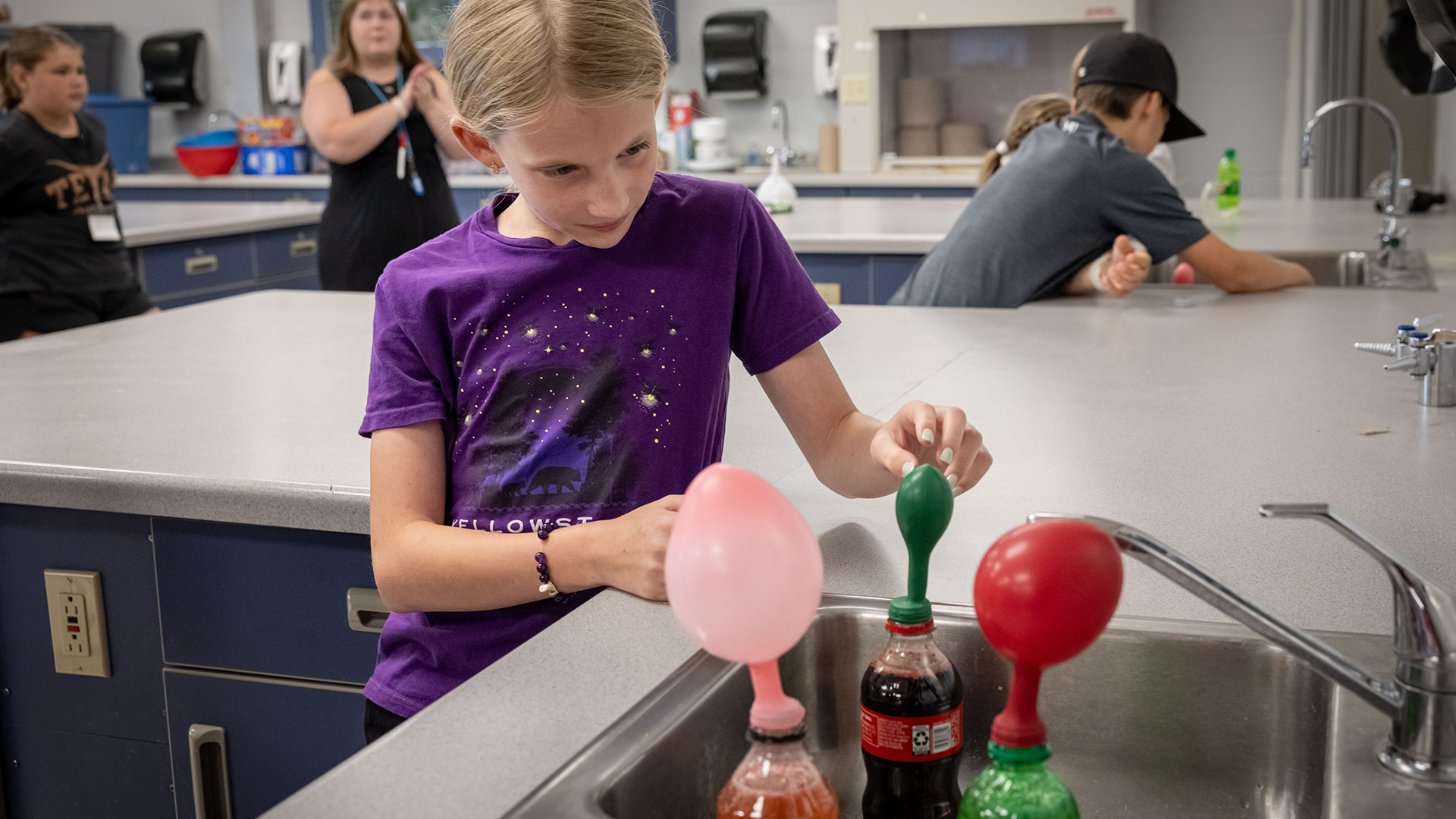 Quinn Kitzman uses her left hand to hold the end of a ballon attached to the mouth of a plastic soda bottle standing next to two other soda bottles topped with inflating balloons in a stainless-steel sink, with a lab bench behind her and other people standing in another lab bench bay and facing another sink, with towel dispensers and storage on the rear wall.