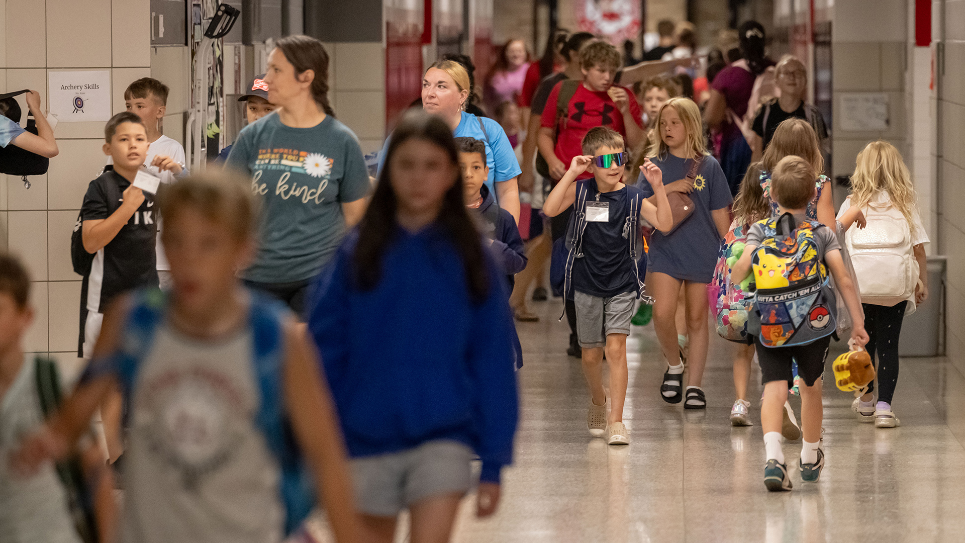 Multiple children in- and out-of-focus walk in both directions along a hallway with vinyl flooring and lockers on either side in the background.