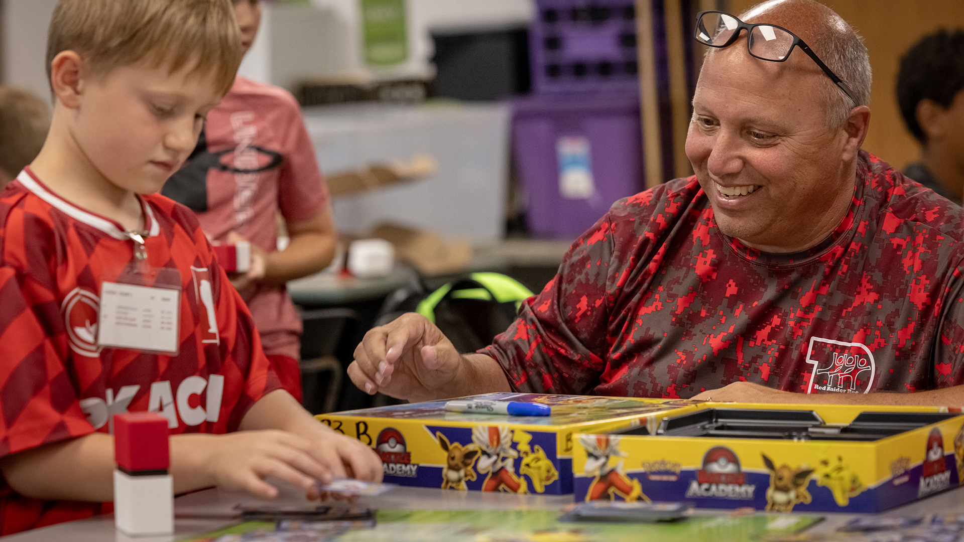 A student plays a board game with Mark Heck on a table, with out-of-focus students and storage bins in the background.