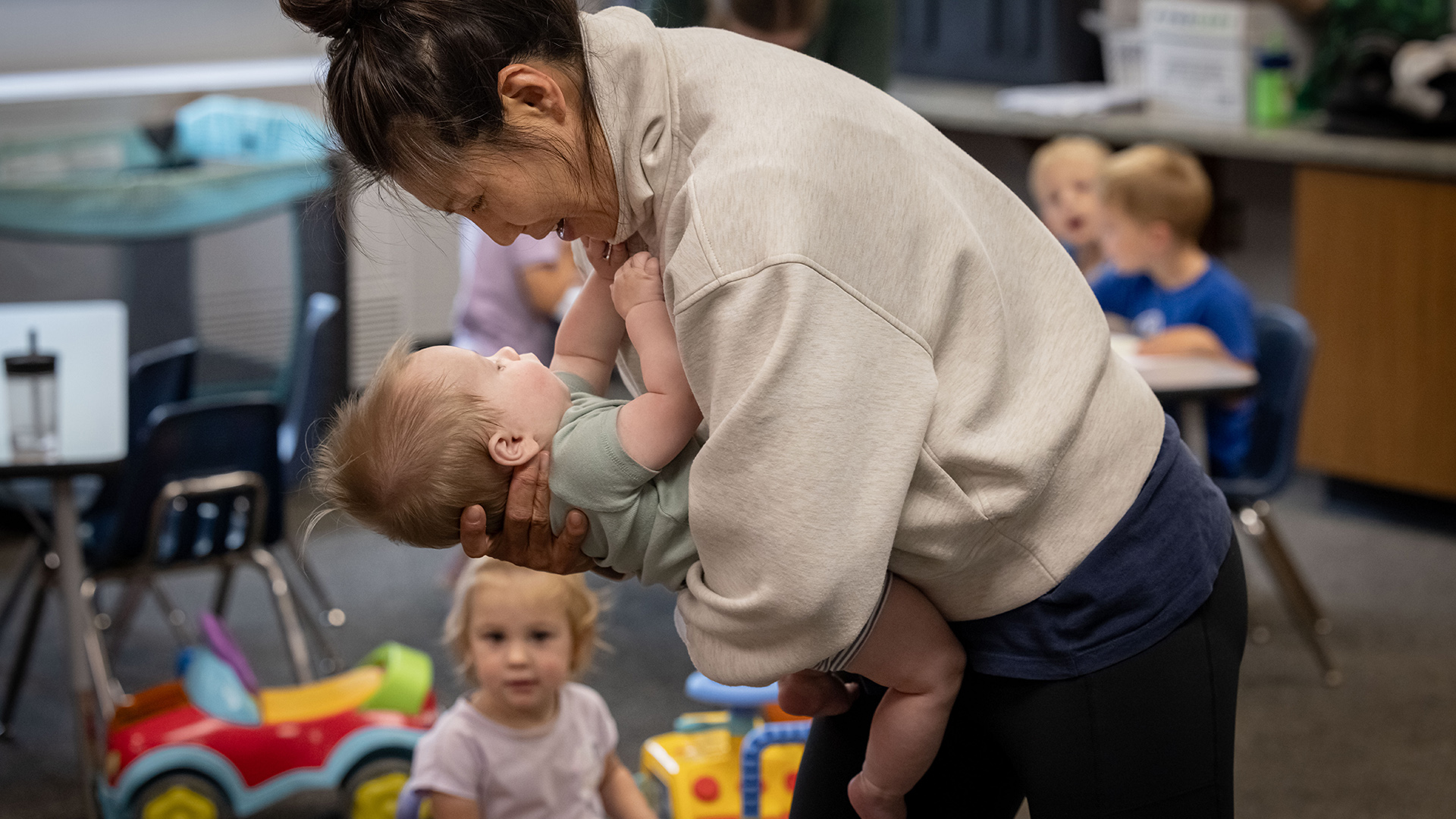 Kyoung Kruger leans forward while holding a toddler in both of her arms while standing in a room with other young children seated on the floor among toy vehicles and in small stacking chairs facing low tables.