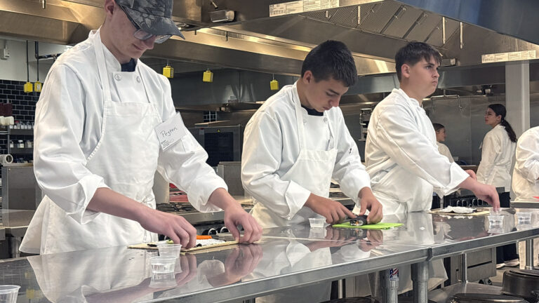 Multiple students wearing chef coats and aprons stand behind a stainless steel table and prepare foods on its surface in a commercial kitchen with tiled walls, large stainless steel appliances and overhead exhaust hoods, with other students standing in the background. Multiple students wearing chef coats and aprons stand behind a stainless steel table and prepare foods on its surface in a commercial kitchen with tiled walls, large stainless steel appliances and overhead exhaust hoods, with other students standing in the background.