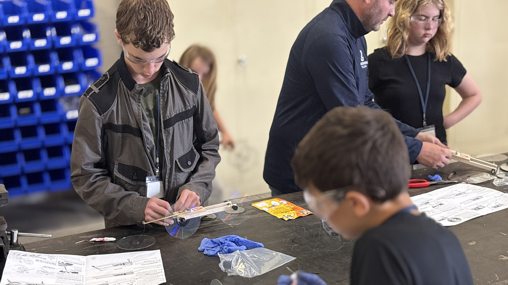 Multiple students and an instructor stand on either side of a wood table and work with different items arrayed on its surface while looking at printed instructions, with an out-of-focus set of plastic storage crates in the background.