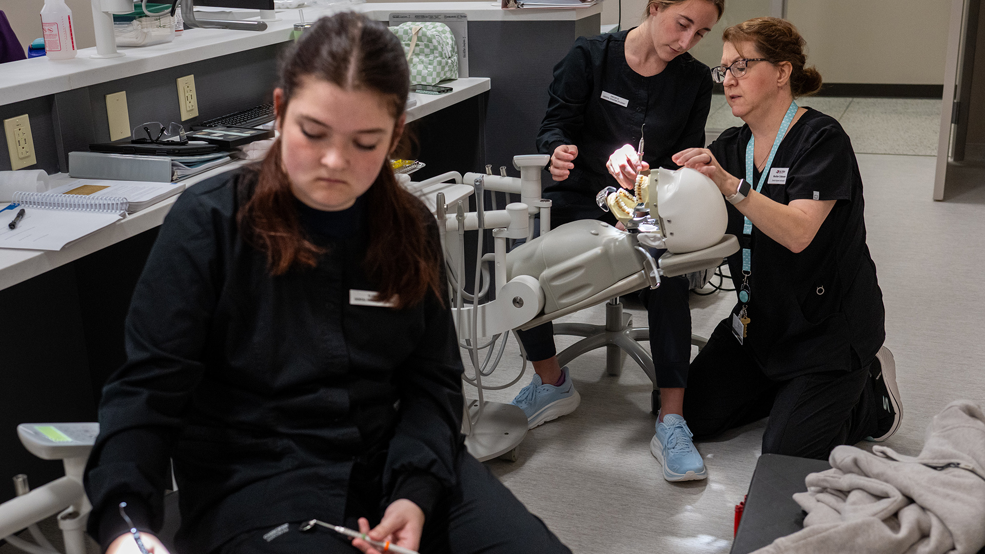 Multiple students sit in rolling chairs and use tools to practice dental work on the teeth of mannequins with open mouths, in a room with multiple rows of desks with computer monitors, water bottles and other items sitting on their surfaces.