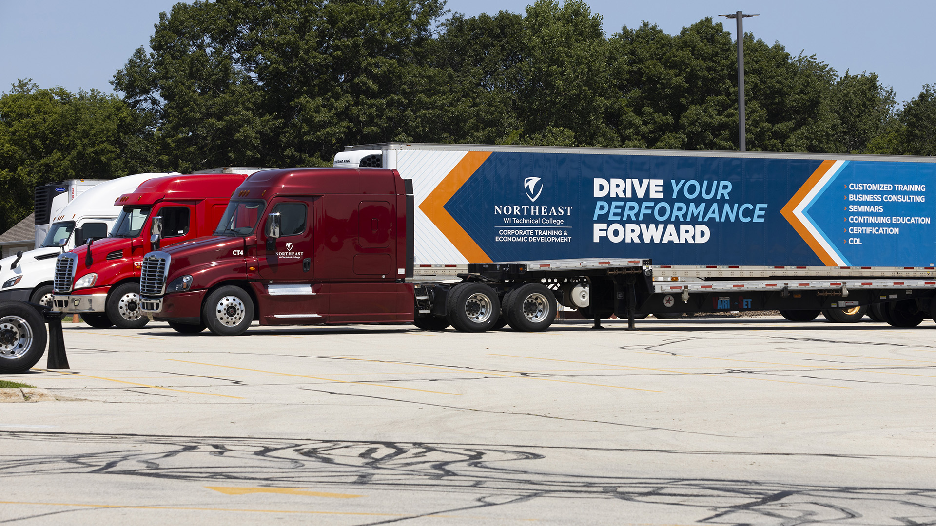 Multiple semi-trucks are parked in a row on a large asphalt parking lot, with the trailer of one with an advertisement for "Northeast WI Technical College" that features the slogan "Drive Your Performance Forward," with multiple trees in the background.