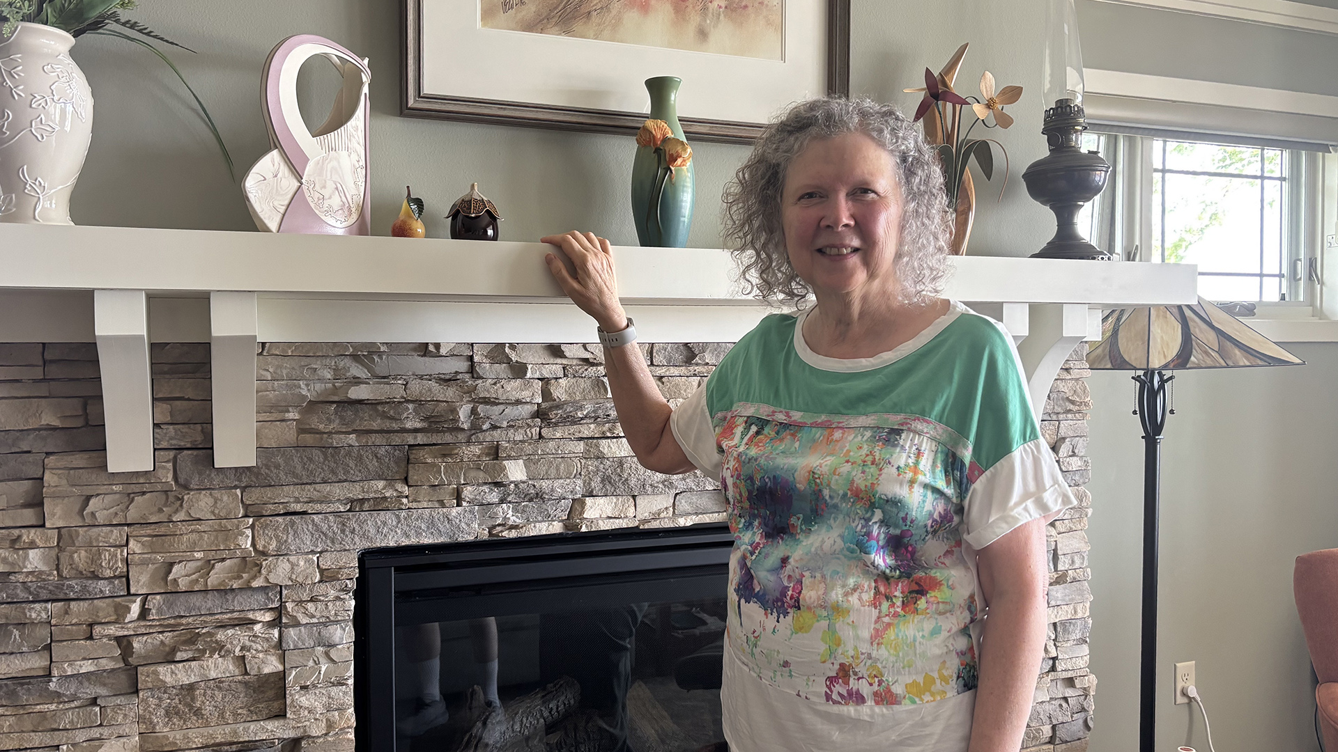 Christine Klotz poses for her portrait while standing in a room in front of a brick fireplace with her right hand on the edge of a mantel with multiple vases on its surface and framed artwork mounted on the wall above, with a lamp in front of a window to the side.