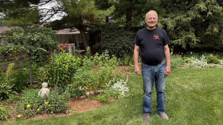 John Short poses for a portrait while standin on a lawn in front areas of garden with different plants surrounded by mulch, with trees and a building in the background. John Short poses for a portrait while standin on a lawn in front areas of garden with different plants surrounded by mulch, with trees and a building in the background.