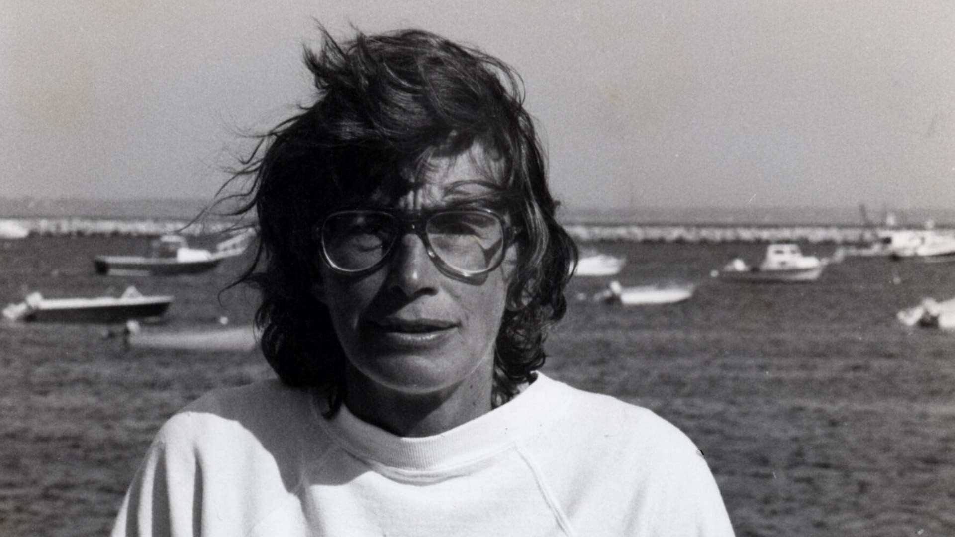 Black-and-white portrait of a young Mary Oliver in glasses and a white sweatshirt, sitting on a dock with boats on the water behind her