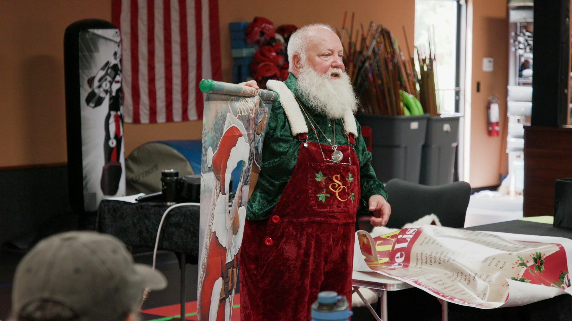 Jeff McMullen, known as Santa Mac, dressed as Santa Claus in a green velvet coat and red apron displays a colorful decorated board in a workshop, creating a festive, cheerful mood.
