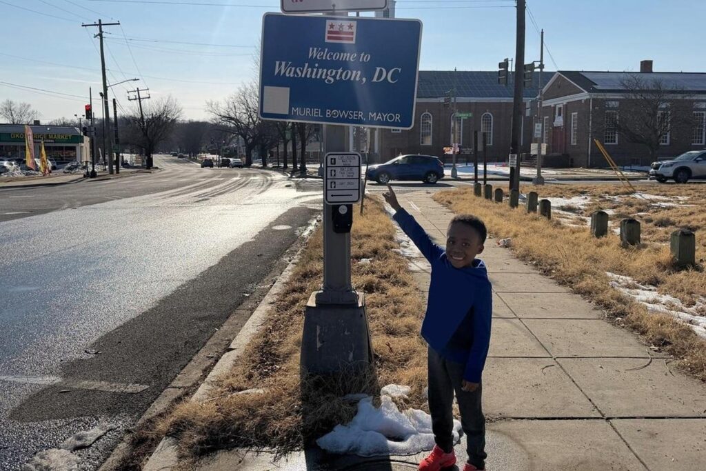 A child on a sidewalk points up to a street sign.