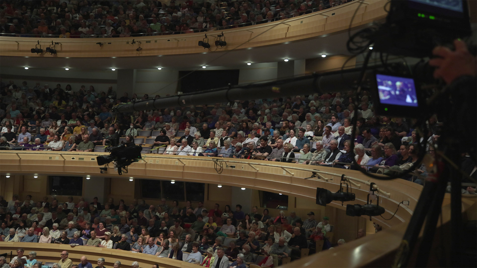 Packed house at the Madison Symphony Orchestra's Centennial Festival Weekend Concert at the Overture Center for the Arts with a diverse audience seated across multiple balconies, while a PBS Wisconsin camera records the lively event.