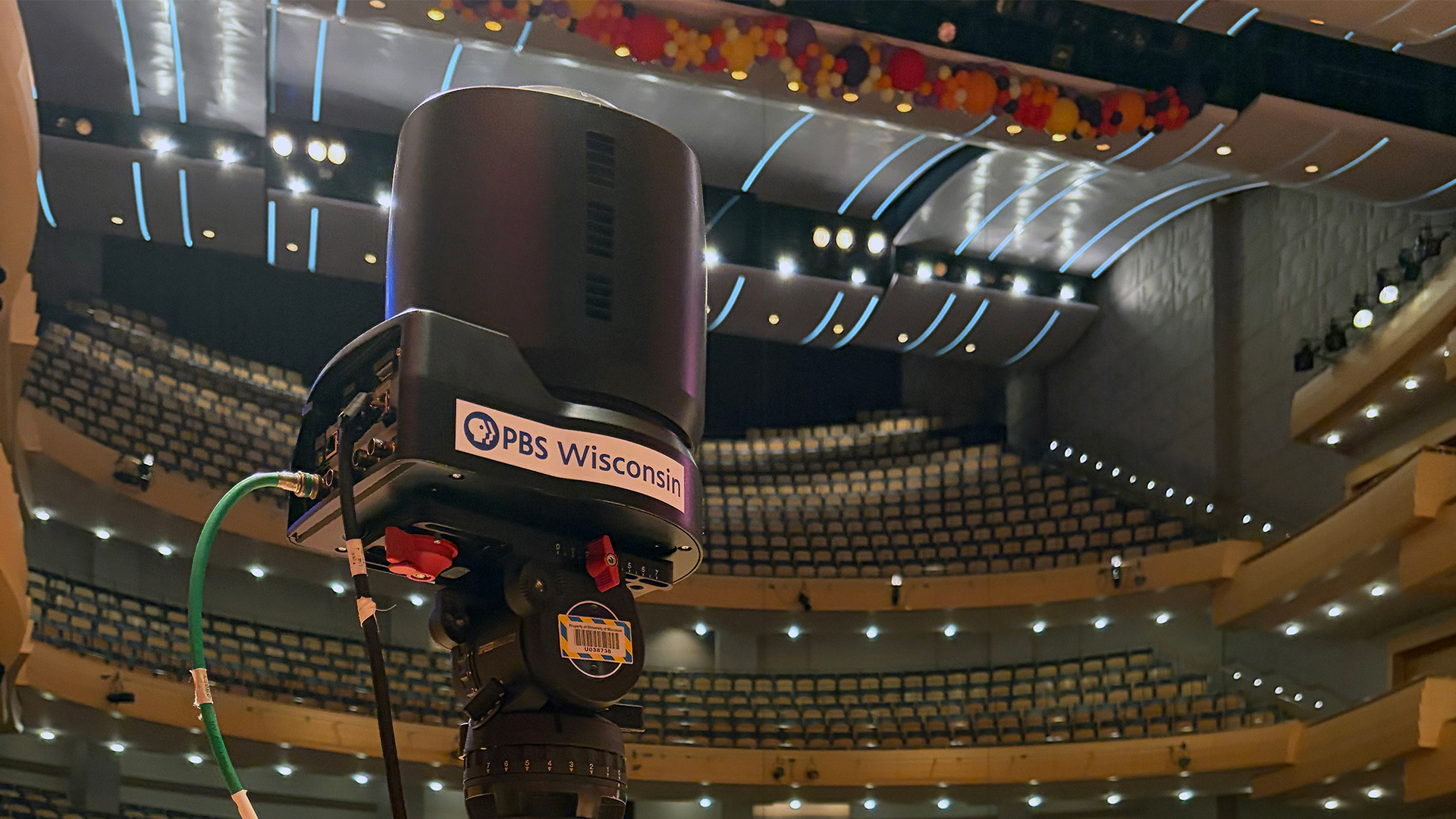 A camera stands on a wooden platform inside the Overture Center for the Fine Arts concert hall, with musicians, crew and empty audience seats visible behind it.