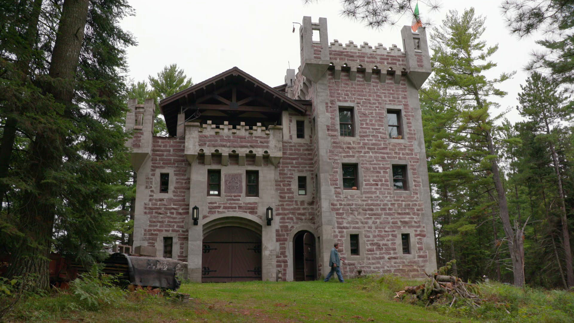 Stone castle with towers, crenellated roofline, arched wooden entrance, and balcony, surrounded by tall trees in a quiet forest.
