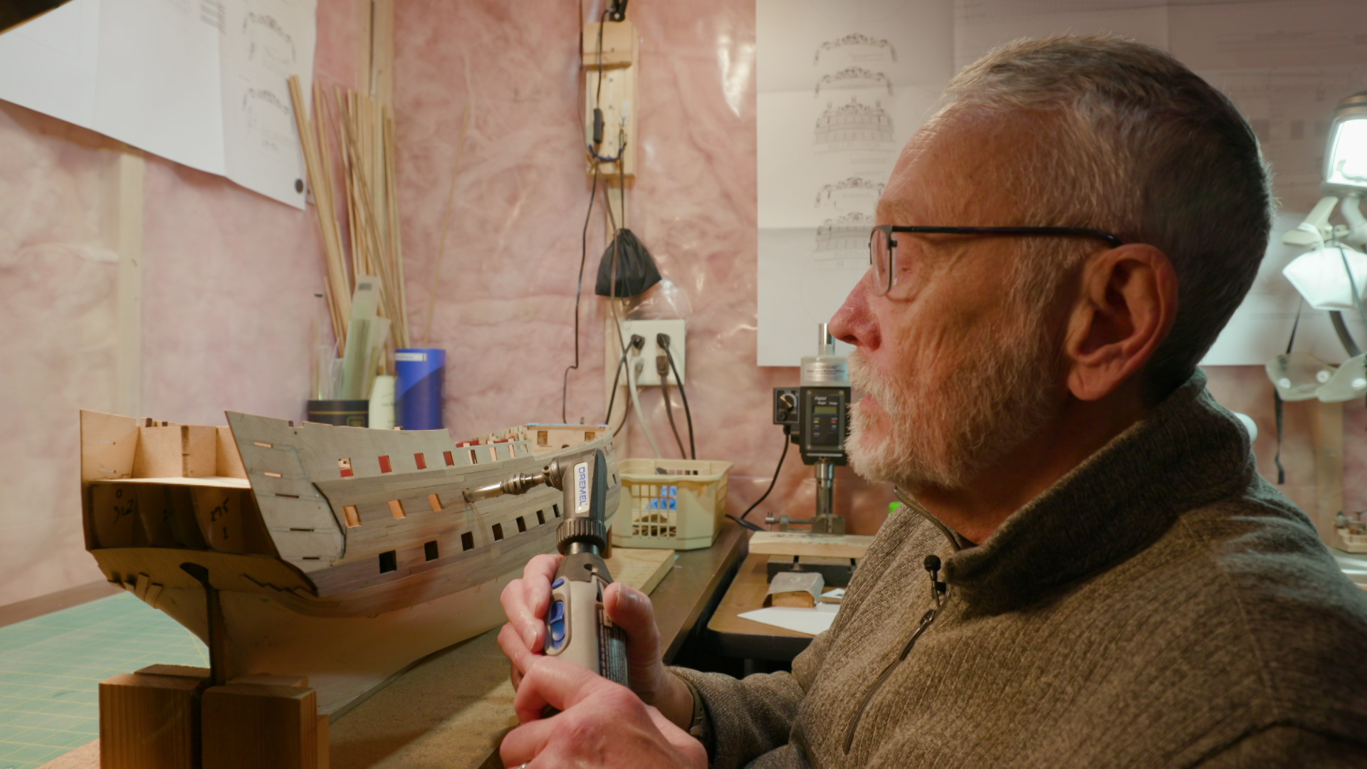 Man with glasses named Scott Safranski examines a detailed model ship on a workbench, holding a tool in a warmly lit workshop; focused, thoughtful mood.