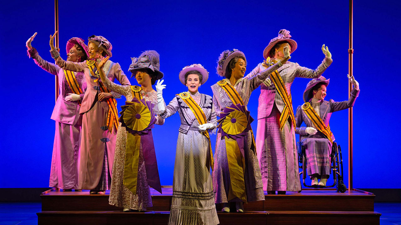 Seven performers in period costumes and suffrage sashes wave onstage; one performer uses a wheelchair.