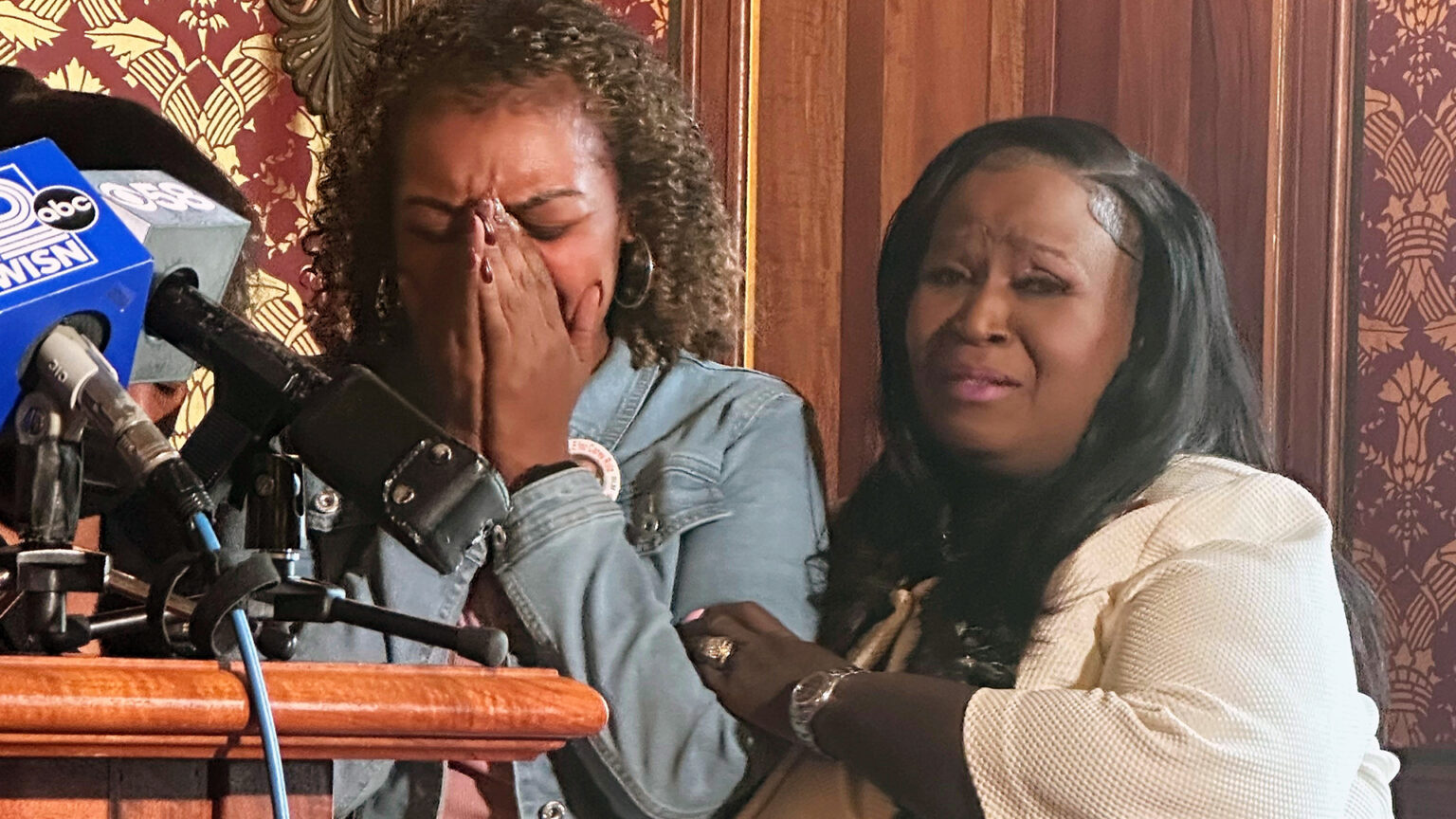 Veronica Ruiz holds her face in her hands and Sheila Stubbs holds Ruiz's left arm, next to a wood podium with multiple microphones with the flags of different media organizations affixed to its surface, in a room with wood wainscoting and toile wallpaper.