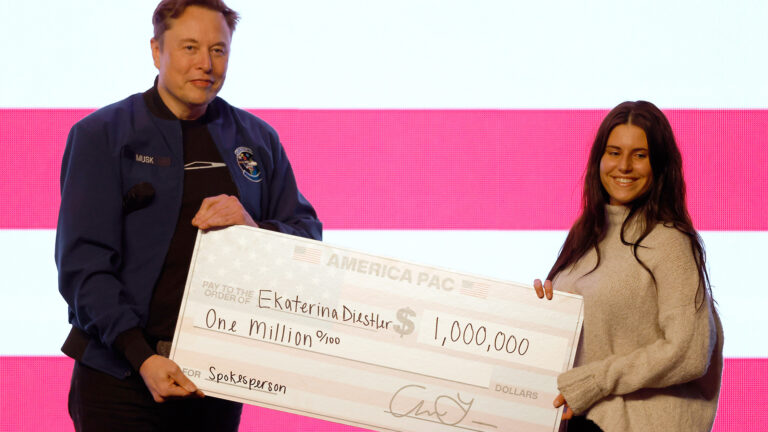 Elon Musk and Ekaterina Diestler stand and hold two ends of an oversized paper check with the words America PAC, pay to the order of Ekaterina Diestler and $1,000,000, with an illuminated screen in the background. Elon Musk and Ekaterina Diestler stand and hold two ends of an oversized paper check with the words America PAC, pay to the order of Ekaterina Diestler and $1,000,000, with an illuminated screen in the background.