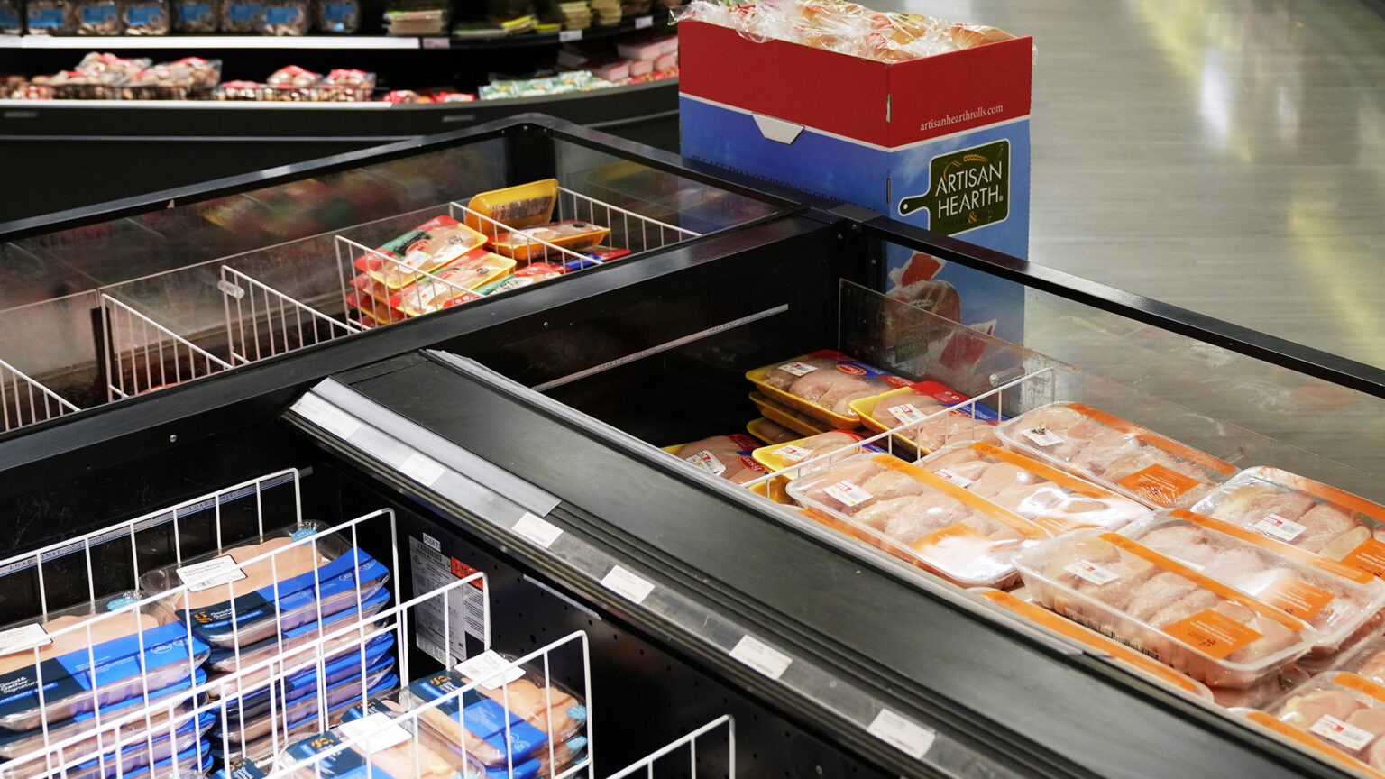 Different poultry cuts and other meat products packaged in trays and encased in plastic wrap sit stacked in three adjacent open-topped coolers, with a cardboard container filled with a bakery product standing next to one, with an out-of-focus open-sided cooler displaying different food products in the background.