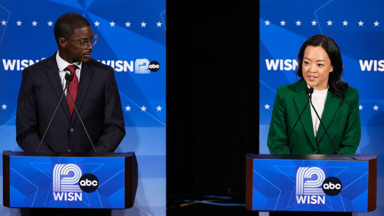 David Crowley looks to his left toward a speaking Francesca Hong, with both standing behind podiums with two microphones affixed to their surface and logos for WISN-TV and ABC on their fronts, and with logo backdrops behind each with rows of five-pointed stars and the WISN 12 and ABC logos.