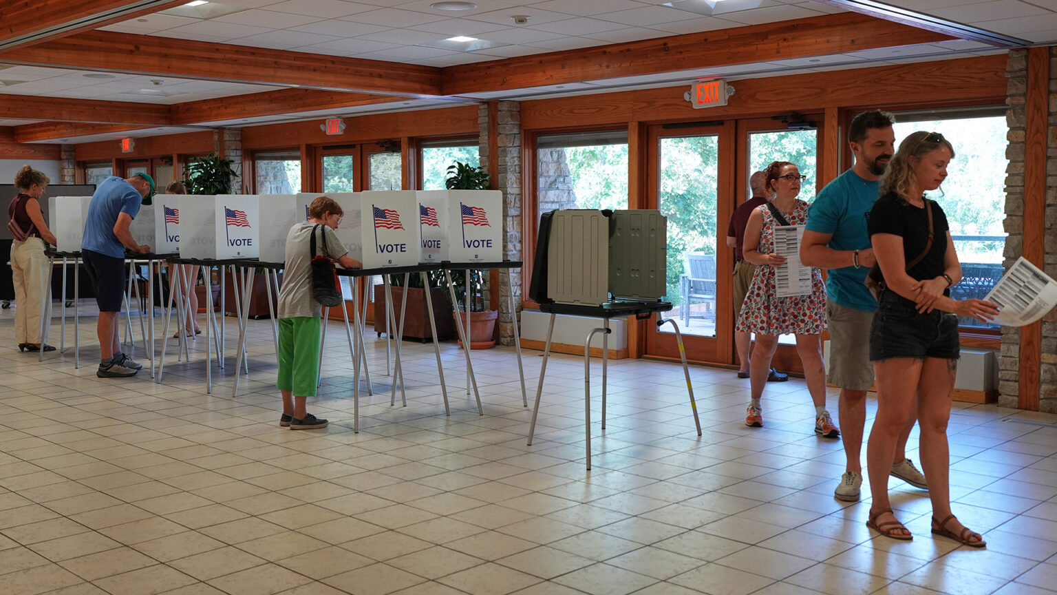 Three people, with two visibly holding paper ballots, stand in a line next to a double row of temporary voting booths with metal legs, plastic trays and cardboard privacy screens with graphics of the U.S. flag and the word Vote on each side, in which several people are facing, in a room with a tile floor and row of wood-trimmed windows and glass doors marked by three illuminated EXIT signs.