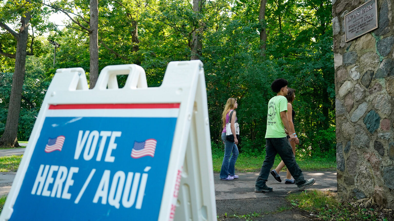 Three people walk along an asphalt-paved path toward the rear side of a corner of a building with stone masonry walls and a bronze plaque reading Lyons Park, with trees in the background and an out-of-focus plastic sandwich board sign with two illustrations of the U.S. flag and the words Vote Here / Aquí on its front face. Three people walk along an asphalt-paved path toward the rear side of a corner of a building with stone masonry walls and a bronze plaque reading Lyons Park, with trees in the background and an out-of-focus plastic sandwich board sign with two illustrations of the U.S. flag and the words Vote Here / Aquí on its front face.