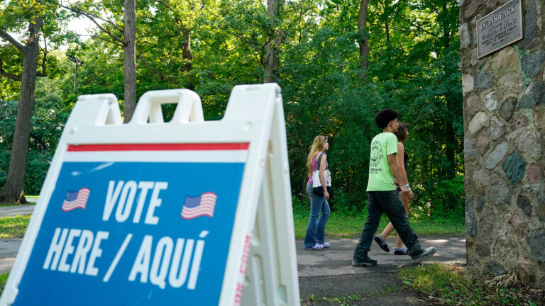 Three people walk along an asphalt-paved path toward the rear side of a corner of a building with stone masonry walls and a bronze plaque reading Lyons Park, with trees in the background and an out-of-focus plastic sandwich board sign with two illustrations of the U.S. flag and the words Vote Here / Aquí on its front face. Three people walk along an asphalt-paved path toward the rear side of a corner of a building with stone masonry walls and a bronze plaque reading Lyons Park, with trees in the background and an out-of-focus plastic sandwich board sign with two illustrations of the U.S. flag and the words Vote Here / Aquí on its front face.