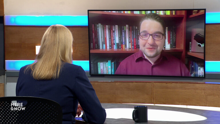 Frederica Freyberg sits at a desk on the Here & Now set and faces a video monitor showing an image of Anthony Chergosky.