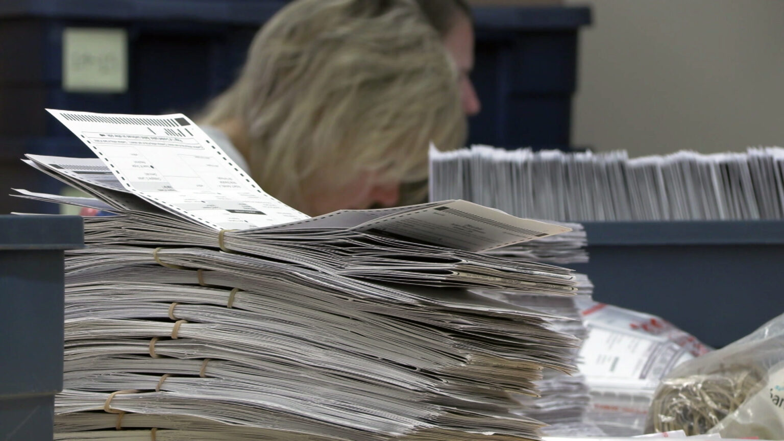 A stack of different sets of absentee ballots bound with rubber bands sits on a table, with out-of-focus people and a tray with absentee ballots arranged on their side in the background. A stack of different sets of absentee ballots bound with rubber bands sits on a table, with out-of-focus people and a tray with absentee ballots arranged on their side in the background.