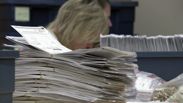 A stack of different sets of absentee ballots bound with rubber bands sits on a table, with out-of-focus people and a tray with absentee ballots arranged on their side in the background. A stack of different sets of absentee ballots bound with rubber bands sits on a table, with out-of-focus people and a tray with absentee ballots arranged on their side in the background.