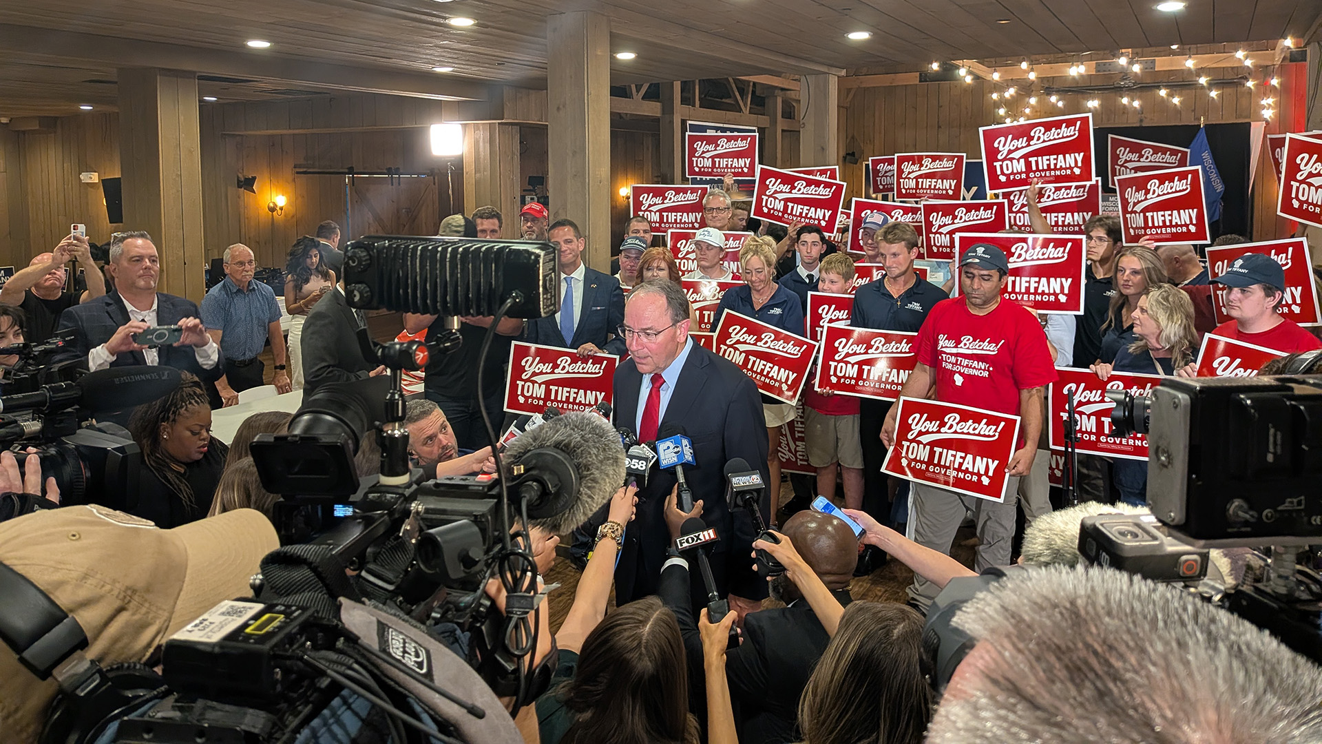 Tom Tiffany speaks while facing a group of reporters in the foreground holding microphones, cameras, smart phones and videocameras, with a group of people holding campaign signs with the words "You Betcha!" and "Tom Tiffany for Governor" standing on risers behind him, in a room with wood beams, wood wall paneling and a wood ceiling.