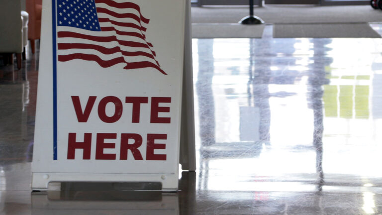 A sandwich board sign with an illustration of a U.S. flag and the words Vote Here stands on a polished vinyl floor reflecting light from windows and doors, with out-of-focus industrial doormats near an entrance in the background. A sandwich board sign with an illustration of a U.S. flag and the words Vote Here stands on a polished vinyl floor reflecting light from windows and doors, with out-of-focus industrial doormats near an entrance in the background.
