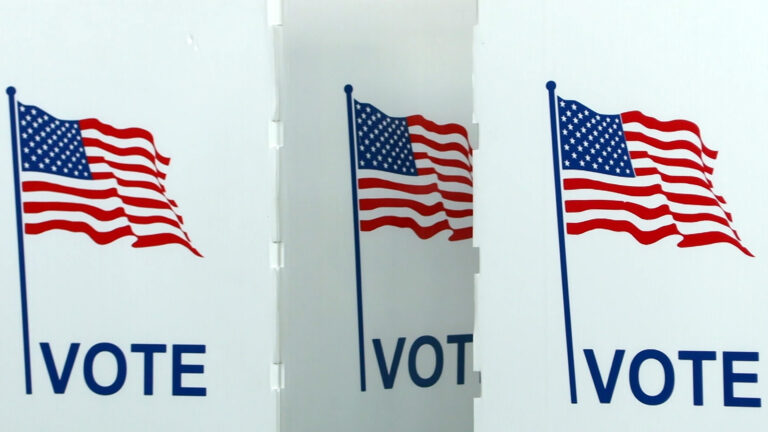 Three plastic wall panels of two voting booths are marked with Illustrations of the U.S. flag and the word Vote, with one obscured by the adjacent booth. Three plastic wall panels of two voting booths are marked with Illustrations of the U.S. flag and the word Vote, with one obscured by the adjacent booth.