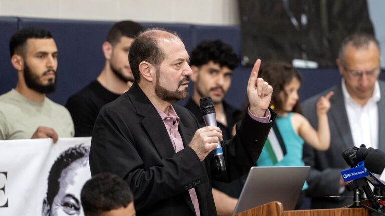Salah Sarsour holds the index finger of his left hand in the air while speaking into a wireless microphone held in his right hand while standing behind a wood podium with a laptop computer on its surface and multiple microphones with the flags of different media organizations affixed to its top, with out-of-focus children standing in the foreground to his side and out-of-focus adults standing behind him in a room with painted concrete block walls. Salah Sarsour holds the index finger of his left hand in the air while speaking into a wireless microphone held in his right hand while standing behind a wood podium with a laptop computer on its surface and multiple microphones with the flags of different media organizations affixed to its top, with out-of-focus children standing in the foreground to his side and out-of-focus adults standing behind him in a room with painted concrete block walls.