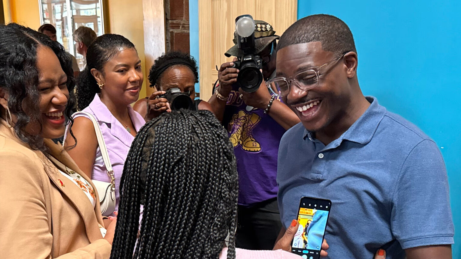 David Crowley smiles while facing a two people who are facing him, one of whom is holding a smart phone in her left hand to record him, with other people standing in the background watching and taking photographs with cameras. David Crowley smiles while facing a two people who are facing him, one of whom is holding a smart phone in her left hand to record him, with other people standing in the background watching and taking photographs with cameras.