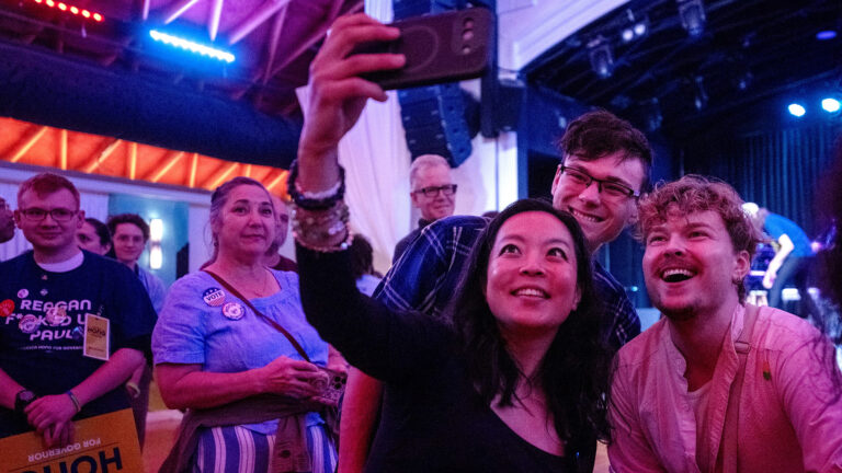 Francesca Hong holds a phone up in her right hand and takes a selfie photo with two people standing behind her, with other people standing in the background of a room with an arched ceiling and a stage at one end with multiple overhead lights.