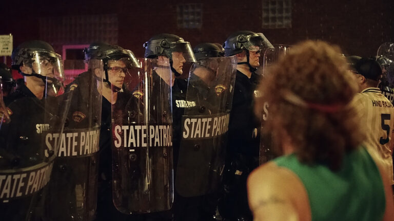 A row of law enforcement officers wearing helmets with face shields and holding transparent riot shields with the words State Patrol across their center span stands and faces multiple people, with a building with glass-block windows in the background.
