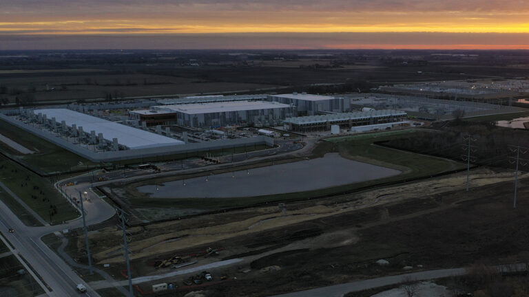 A multi-color partly cloudy sky casts shadows over a landscape of multiple low, large buildings surrounded by roads, retention ponds, power line poles and fields. A multi-color partly cloudy sky casts shadows over a landscape of multiple low, large buildings surrounded by roads, retention ponds, power line poles and fields.