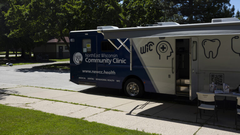 A recreational vehicle with the words NorthEast Wisconsin Community Clinic, the URL www.newcc.health and different graphics is parked on the side of a road next to a sidewalk, with its open side awning providing shade to a folding table and chairs in place next to and open door, with trees and a building in the background. A recreational vehicle with the words NorthEast Wisconsin Community Clinic, the URL www.newcc.health and different graphics is parked on the side of a road next to a sidewalk, with its open side awning providing shade to a folding table and chairs in place next to and open door, with trees and a building in the background.