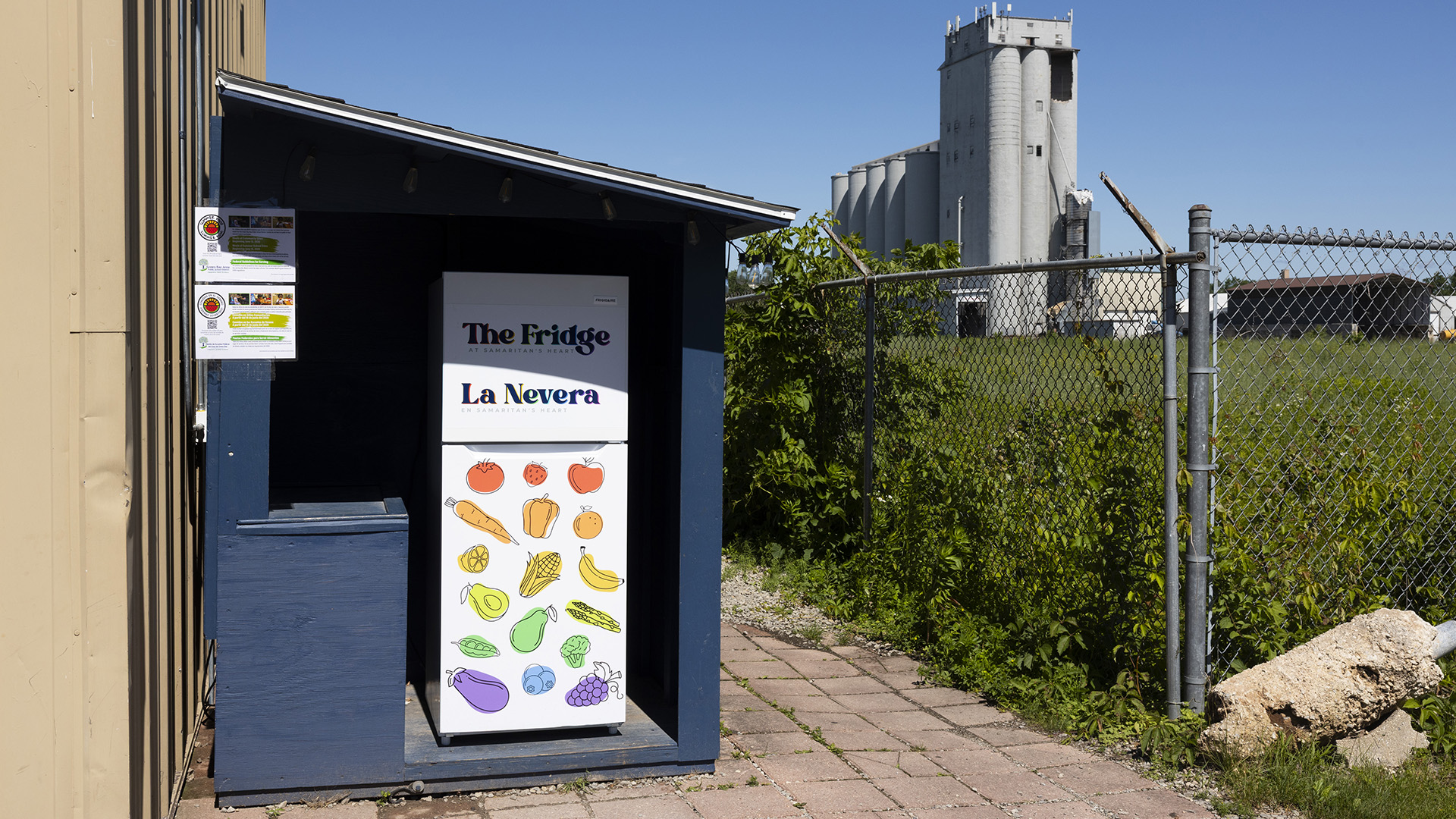 A refrigerator with labels on its upper freezer door reading "The Fridge" and "Le Nevera" and with decals of fruits and vegetables on its lower cooler door stands in an open lean-to style shed next to a building on a paved patio, with a chainlink fence topped by barbed wire standing on the other side containing a field with tall prairie grasses in front of a grain elevator and other buildings in the background.