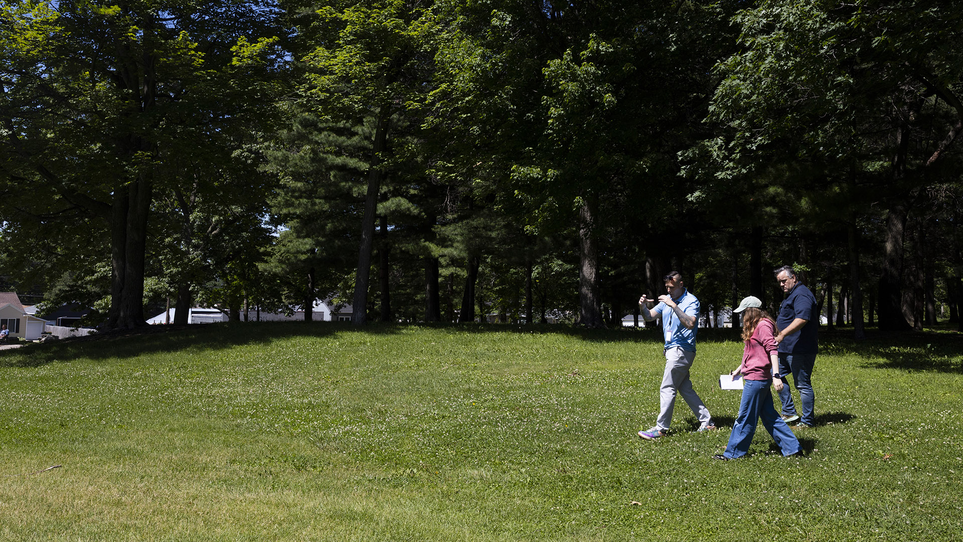 Peter Silski, Astrid Code and Jeremy Pingel walk along a lawn in front of a stand of trees, with low buildings behind them in the background.