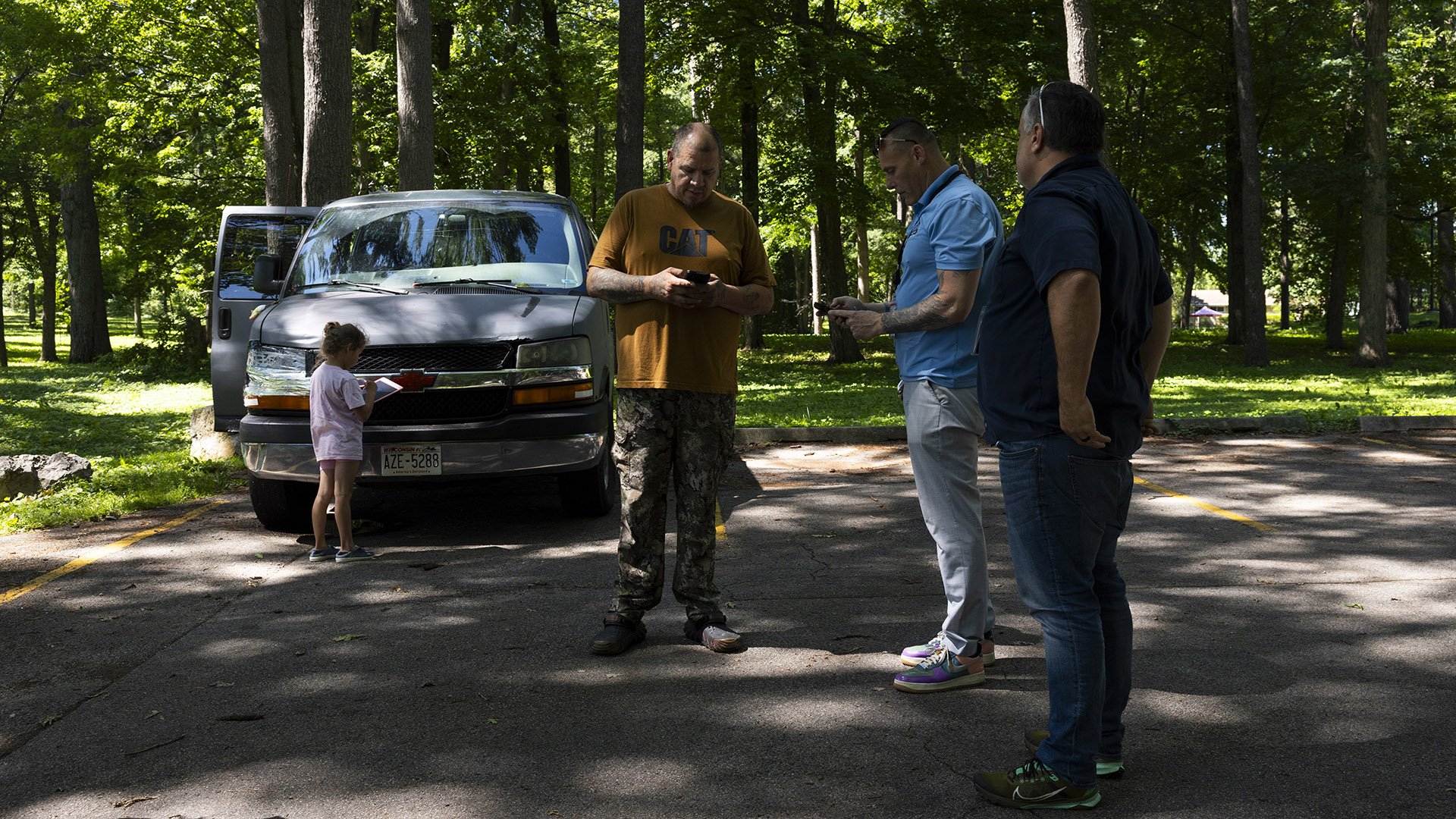 James Gray stands and faces Peter Silski, who is standing next to Jeremy Pingel, with a child standing in front of a parked pickup truck with an open passenger-side door in the background, in a parking lot shaded by trees canopy, with numerous trees standing over a lawn in the background.