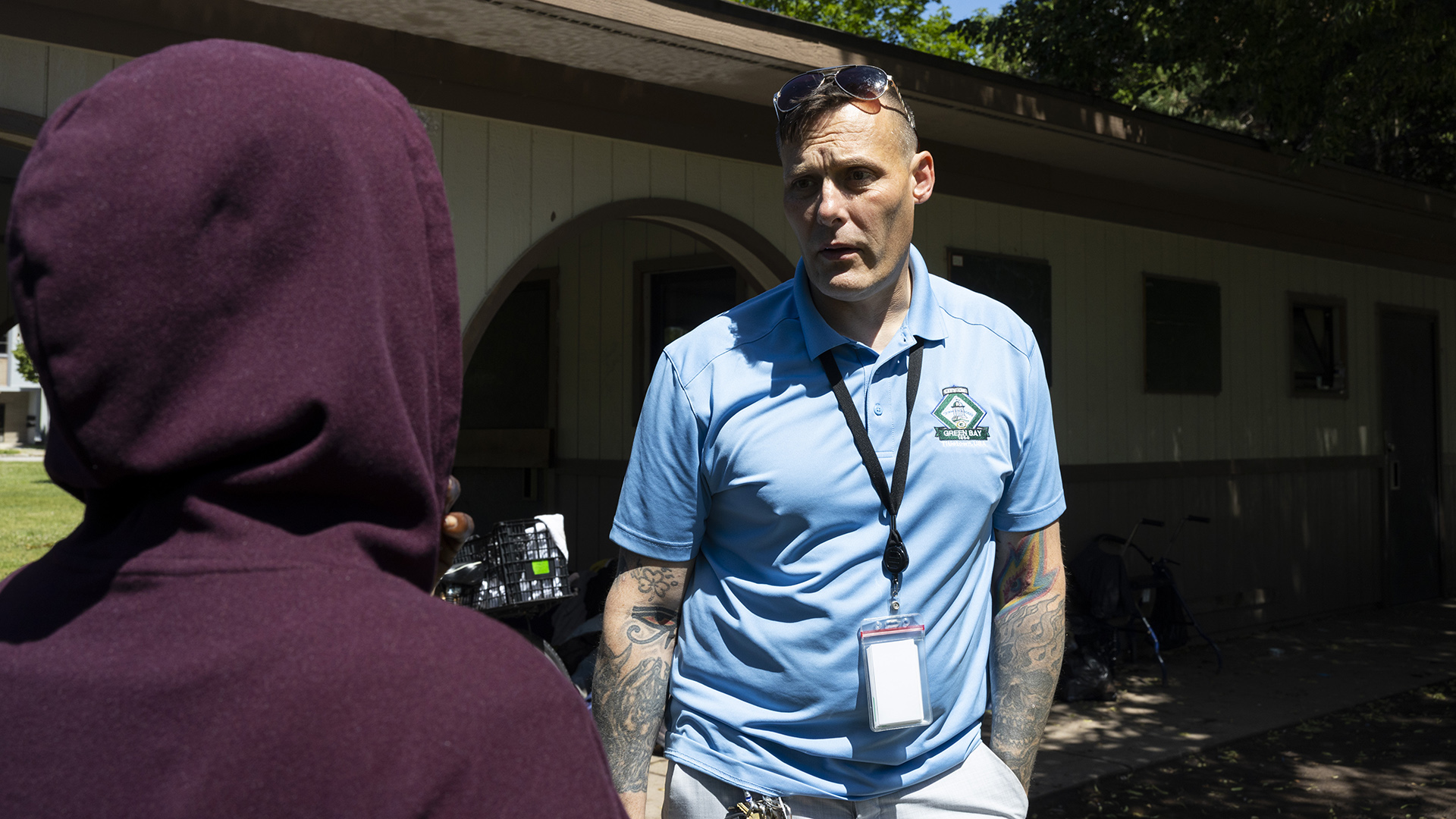 Peter Silski stands and talks to a person in the foreground who is wearing a hoodie over their head and is facing him, with a single-story building with an arched entrance and multiple windows shaded by adjacent trees in the background.