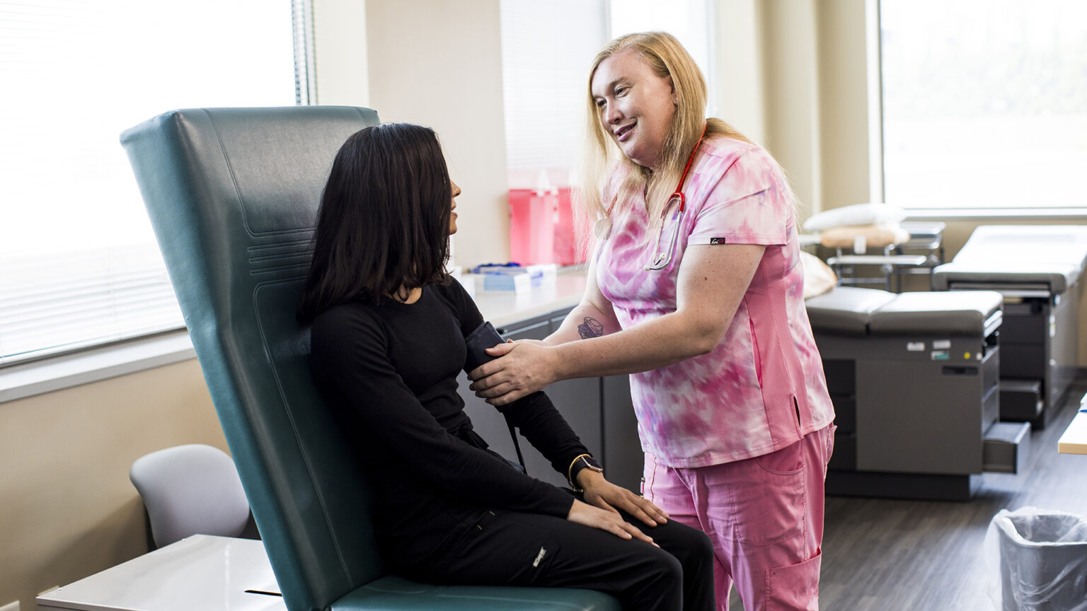 Violet LaClair attaches a blood pressure monitor cuff to the arm of a person seated in a high-backed exam chair, in a room with medical exam tables and cabinets along a wall in front of windows with light streaming through blinds. Violet LaClair attaches a blood pressure monitor cuff to the arm of a person seated in a high-backed exam chair, in a room with medical exam tables and cabinets along a wall in front of windows with light streaming through blinds.