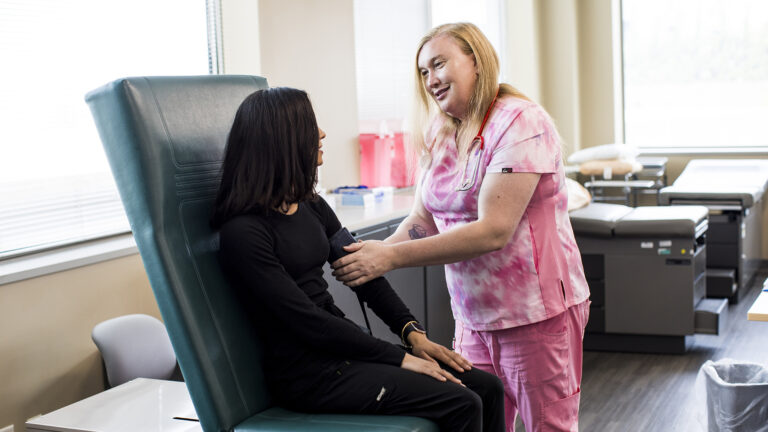 Violet LaClair attaches a blood pressure monitor cuff to the arm of a person seated in a high-backed exam chair, in a room with medical exam tables and cabinets along a wall in front of windows with light streaming through blinds. Violet LaClair attaches a blood pressure monitor cuff to the arm of a person seated in a high-backed exam chair, in a room with medical exam tables and cabinets along a wall in front of windows with light streaming through blinds.